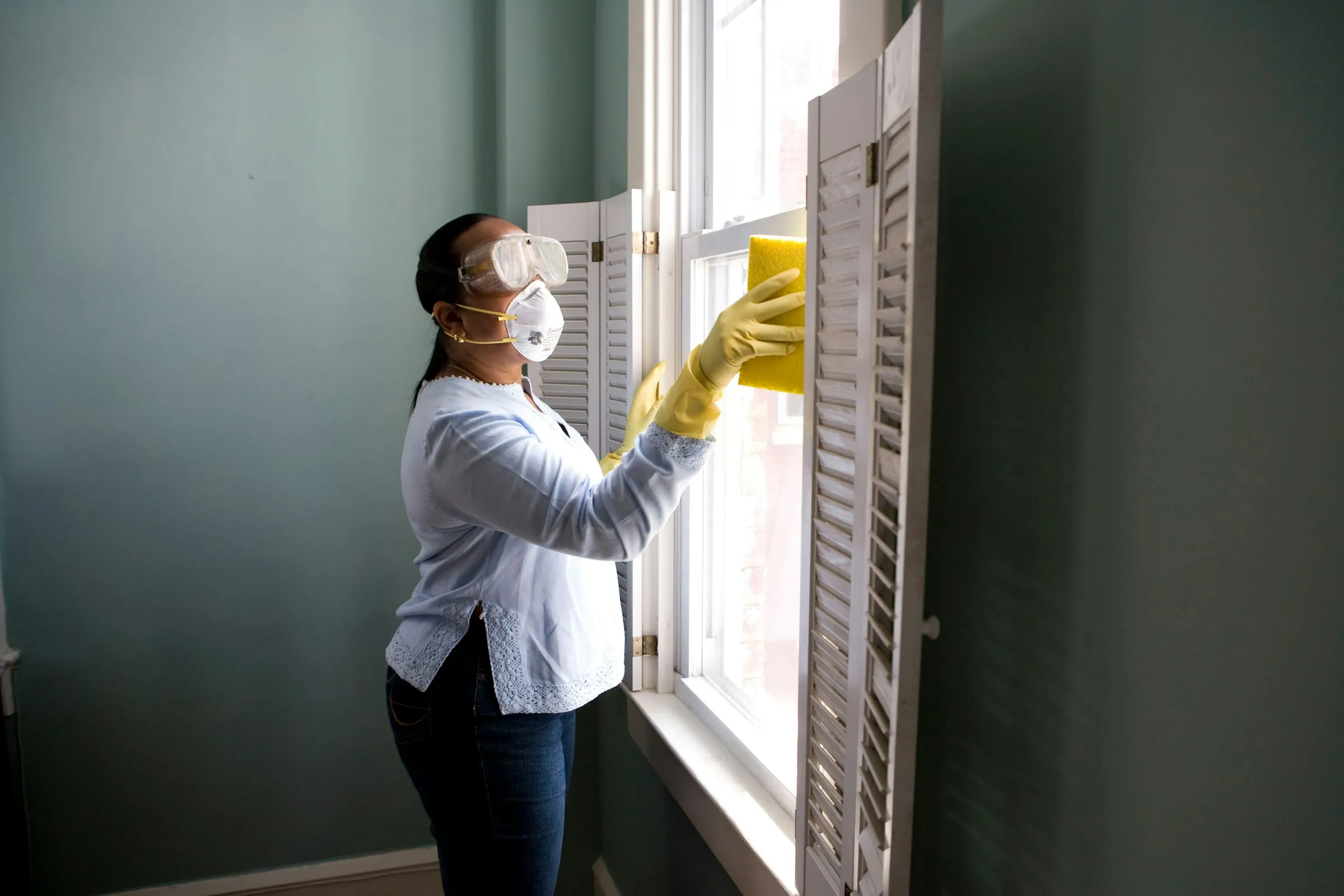 A cleaner in gloves and a mask wiping the frame of a window with white shutters