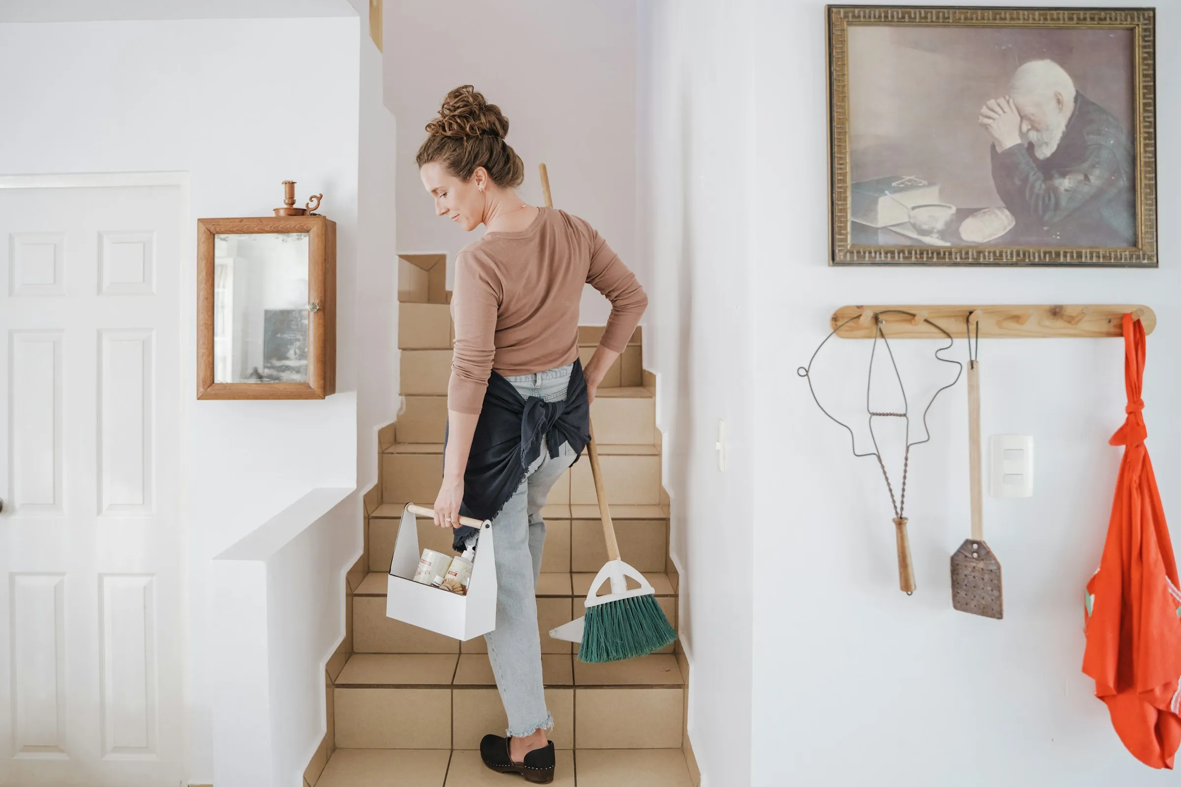 A cleaner carrying a cleaning caddy and a broom up the stairs of a bright home
